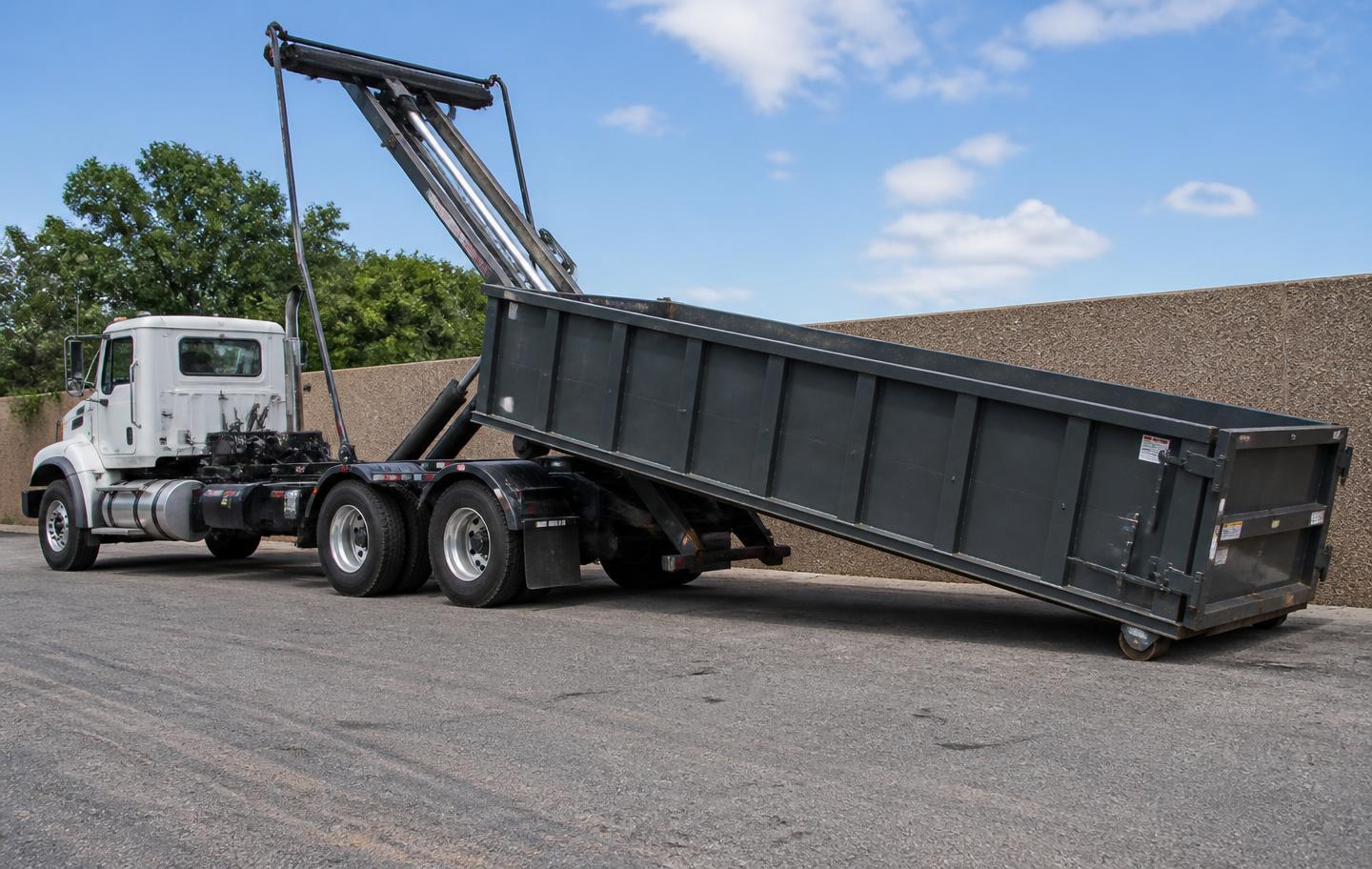 Roll-off truck tilting its bed to slide an open-top steel container onto the pavement
